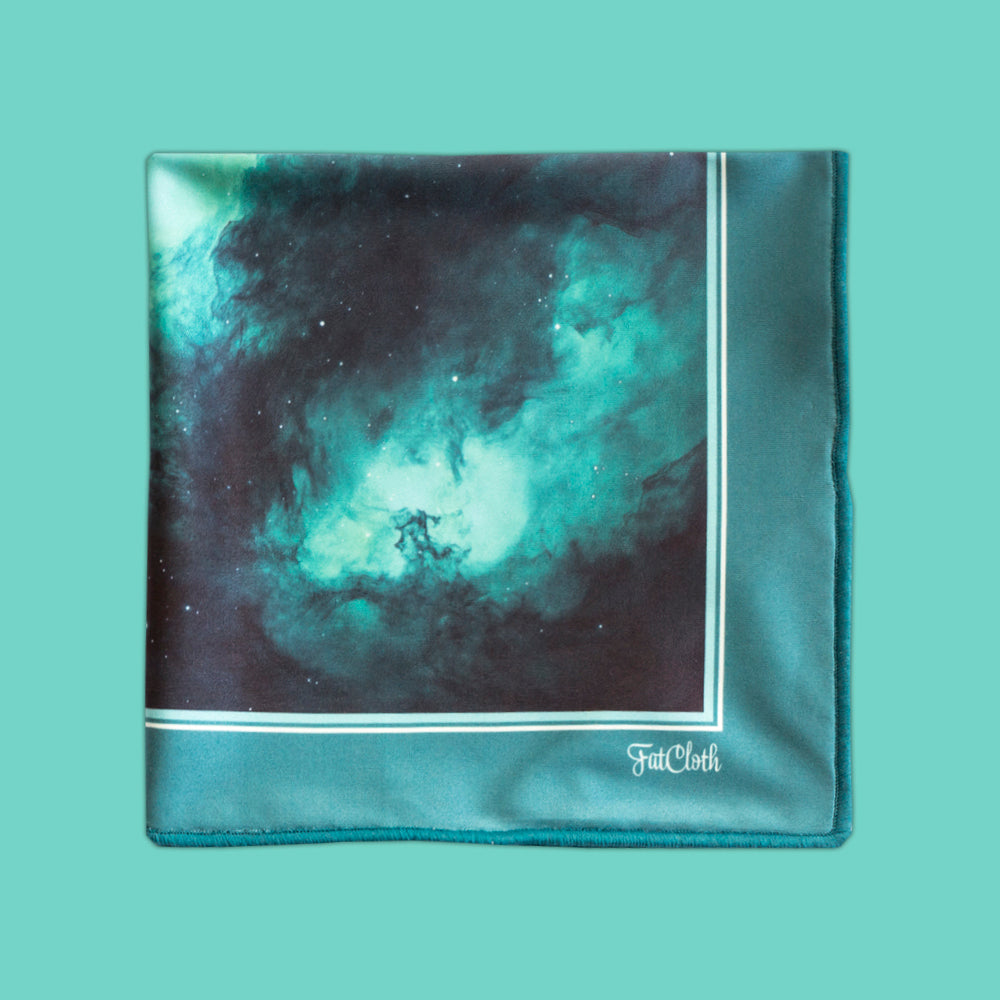 A multipurpose pocket square with a printed nebula design in teal, black, and white, finished with a solid teal border and a white FatCloth logo on the bottom right, set against a solid teal background.
