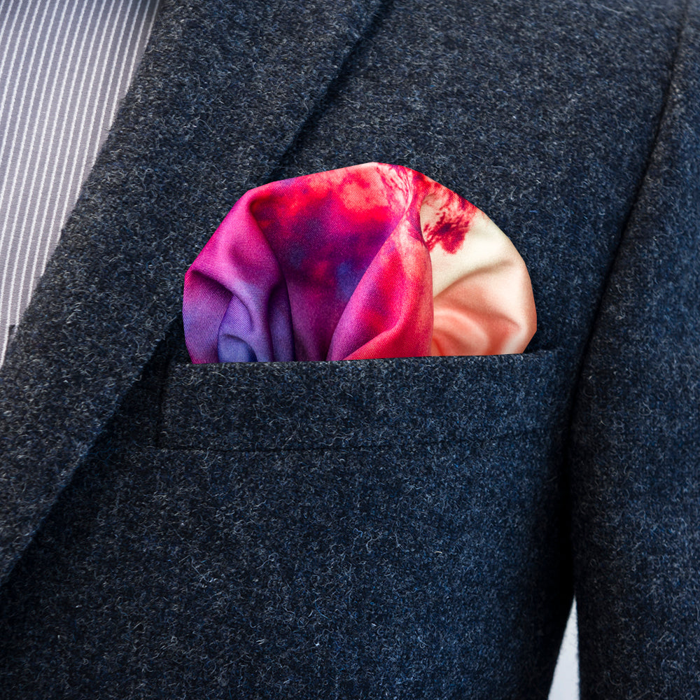 A close-up of a dark gray textured suit jacket with a colorful nebula pocket square tucked into the breast pocket. The pocket square features a vibrant pattern of magenta, purple, and yellow.
