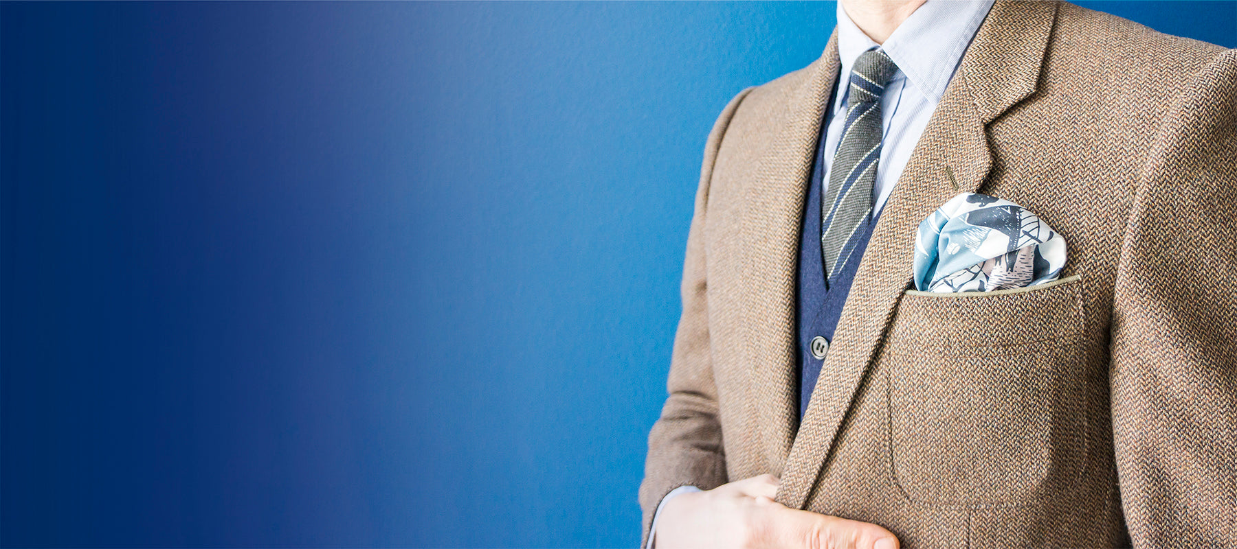 Man wearing light blue pocket square in the pocket of a brown tweed blazer with gradient blue background