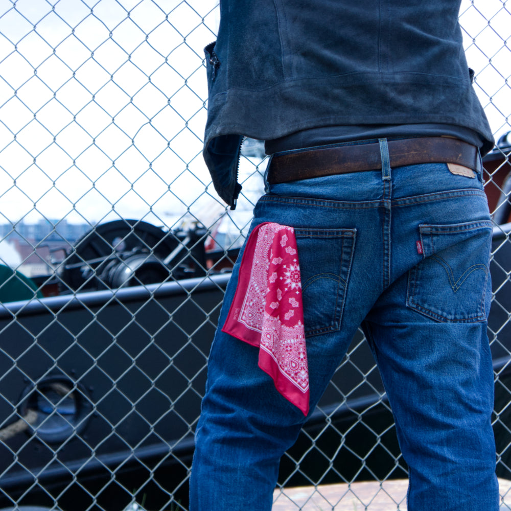 Person wearing blue jeans with a red bandana in left rear pocket, standing in front of a chain-link fence.