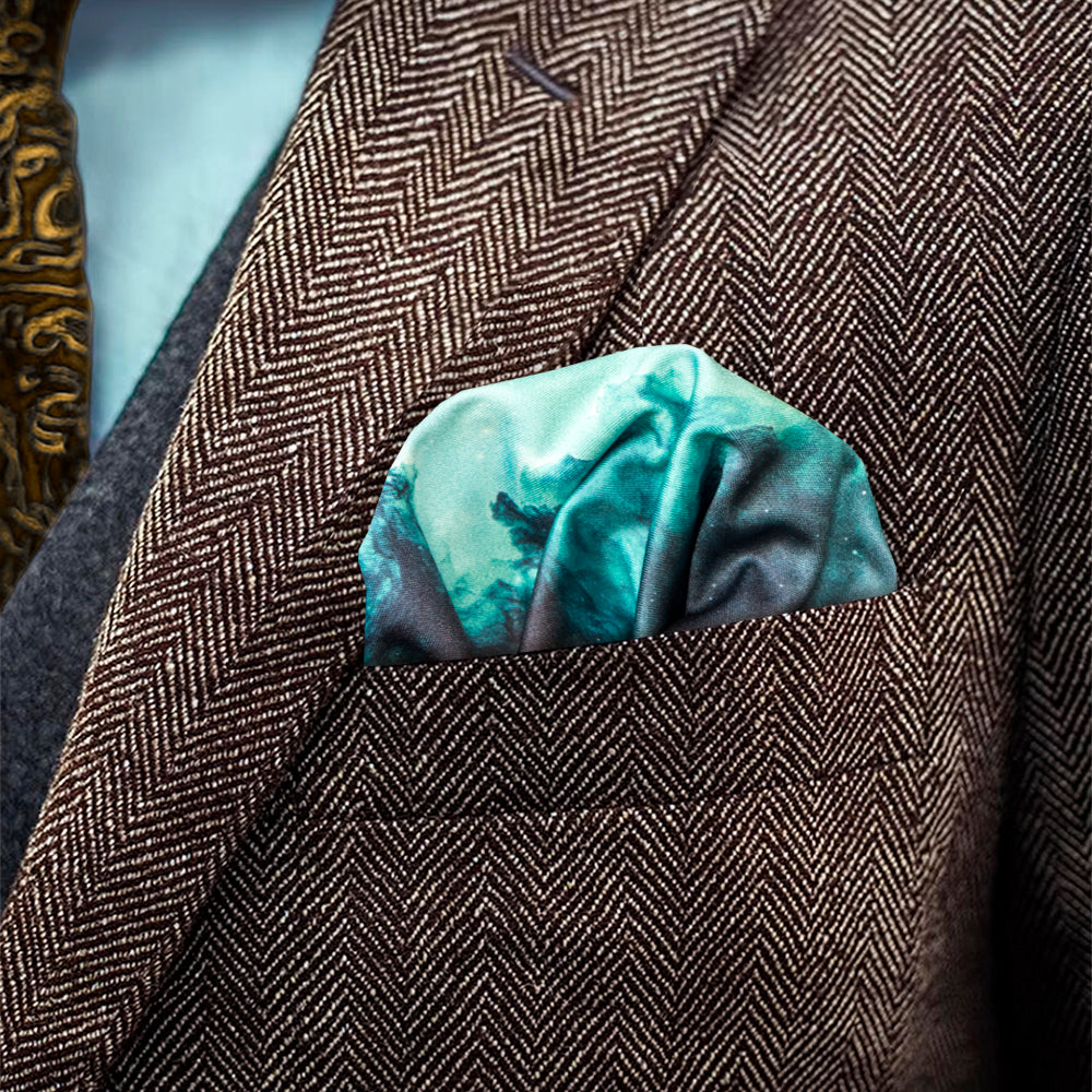 A close-up of a brown herringbone blazer jacket with a puff folded pocket square in the chest pocket, featuring a teal and dark gray nebula pattern.