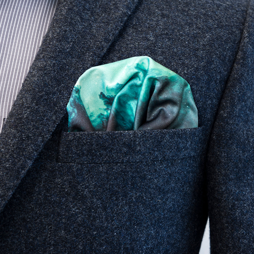 A close-up of a textured gray suit jacket with a FatCloth pocket square tucked into the breast pocket. The pocket square has a nebula gradient pattern in shades of teal, green, and dark gray.