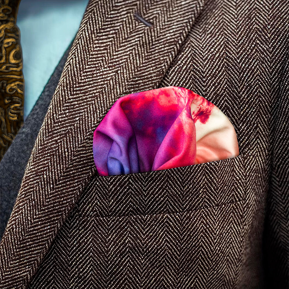 A close-up of a brown herringbone blazer jacket with a colorful, puff folded pocket square tucked into the breast pocket. The pocket square features a vibrant gradient pattern in shades of pink, purple, yellow and blue.